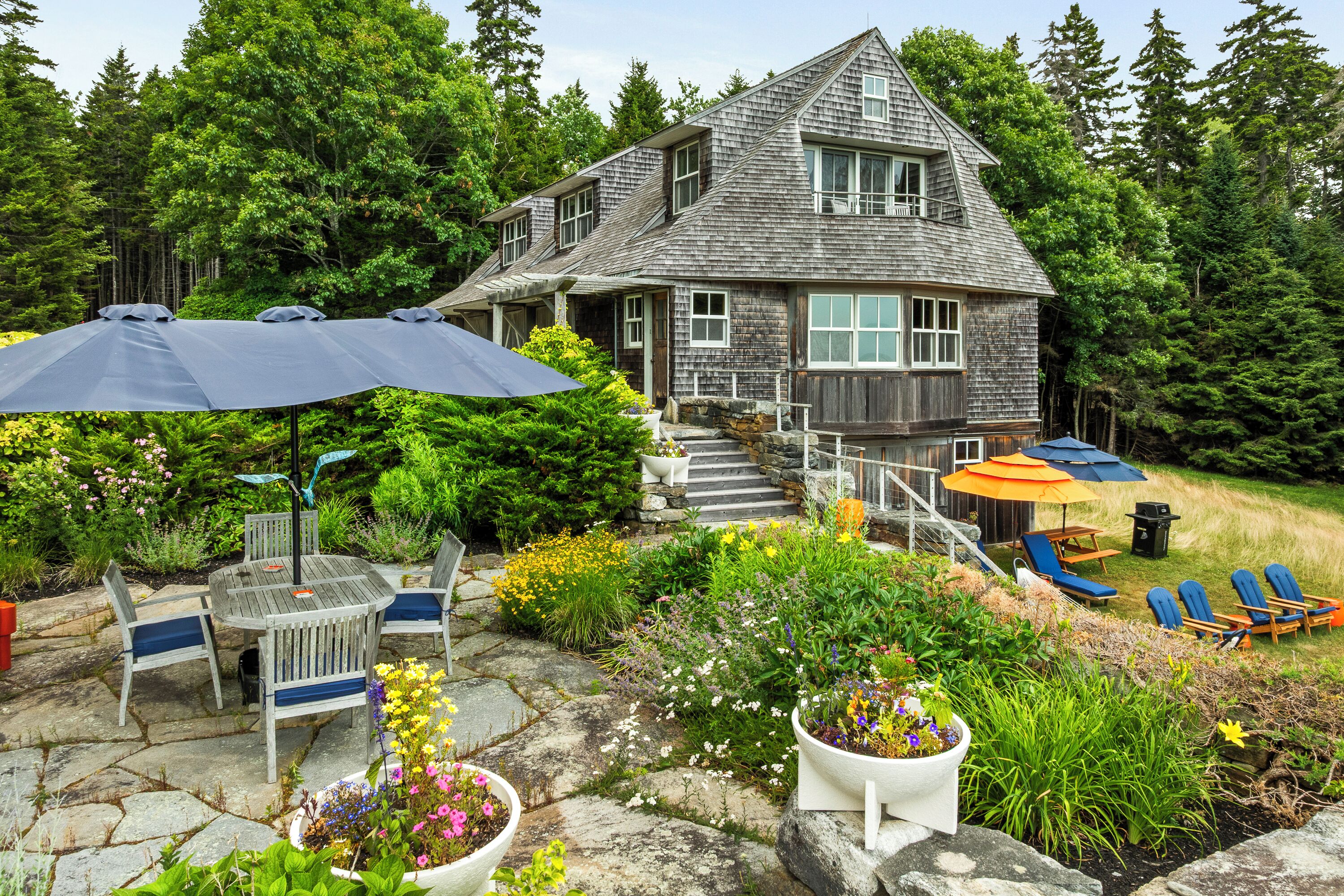 A two-story wooden house with a stone staircase leading to the entrance, surrounded by lush greenery and a patio area with outdoor furniture.
