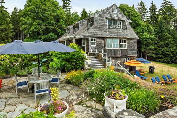 A two-story wooden house with a stone staircase leading to the entrance, surrounded by lush greenery and a patio area with outdoor furniture. - NEW! Quaint 2BR apt in Harpswell with private beach area and beautiful gardens (Harpswell)