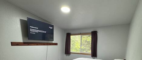 A bedroom with a bed, a TV mounted on the wall, a window with curtains, and a bedside table with a lamp.