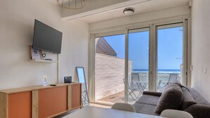 A modern room with a flat-screen TV mounted on the wall, a wooden cabinet, a white table, and a sofa with a view of the beach through large sliding glass doors. - Le Mosala 1 - Climatisation - Vue mer (Saint-Leu)
