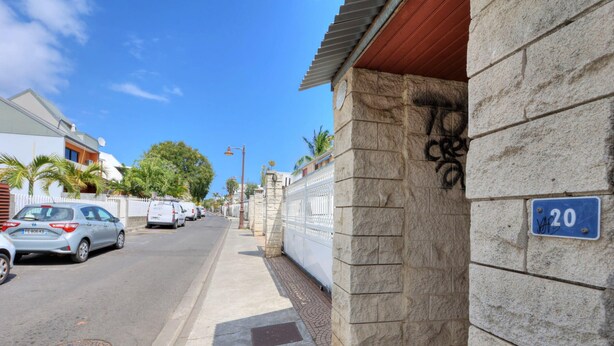 A street view with parked cars, a sidewalk, and a building with a stone facade and a blue sign indicating the address. - Le Mosala 1 - Climatisation - Vue mer (Saint-Leu)