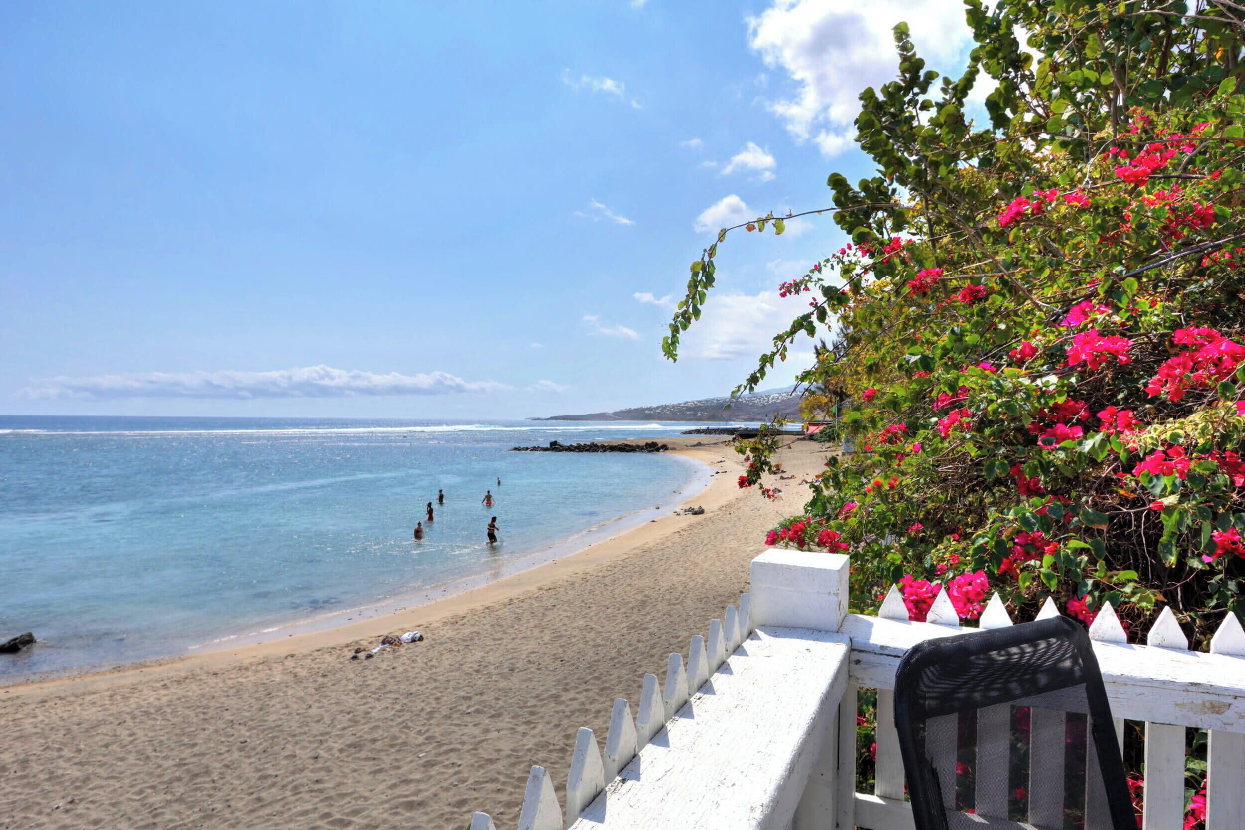 A beach with clear blue water, a sandy shore, and a white fence with a chair.