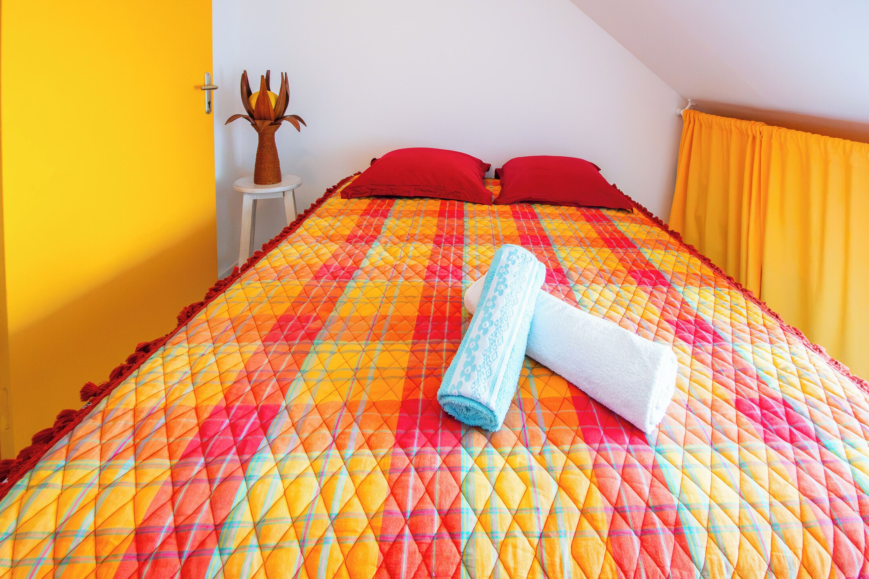 A bed with a colorful quilt, red pillows, and a rolled towel.