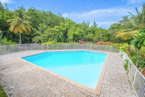 Ein rechteckiger Pool mit Bordstein aus Steinplatten, umgeben von einem weißen Metallzaun, vor einer Kulisse aus üppigem Grün und Bäumen.