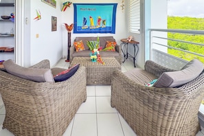 A cozy living area with wicker furniture, a colorful painting of sailboats, and a view of greenery outside. - Le Nid bleu -Climatisation -Proche plage (SAINTE ANNE)