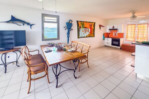 A dining area with a wooden table and chairs, a window with a view of the sea, and a kitchen with white cabinets and a red and yellow tiled backsplash. - Le Nid bleu -Climatisation -Proche plage (SAINTE ANNE)
