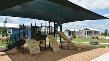 Un parque infantil con estructuras para escalar y toboganes, rodeado de mesas de picnic y cubierto por una sombra.