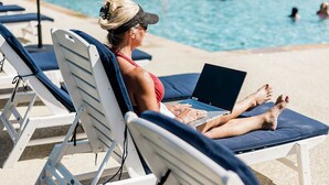 Eine Person benutzt einen Laptop auf einer Liege am Pool.