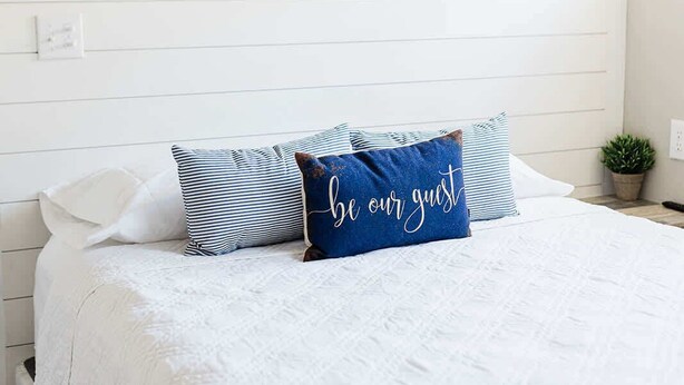 Ein Schlafzimmer mit weißer Bettdecke, einem blauen Kopfkissen mit der Aufschrift 'be our guest' und einer weißen Klimaanlage an der Wand.
