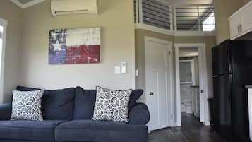 A living room with a dark sofa, patterned pillows, a wall-mounted air conditioner, and a kitchen area with a black refrigerator.