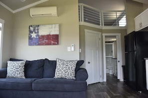 A living room with a dark sofa, patterned pillows, a wall-mounted air conditioner, and a kitchen area with a black refrigerator.