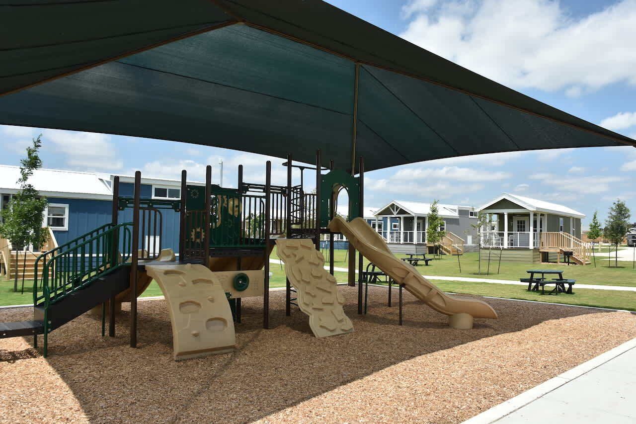 Ein Spielplatz mit Klettergerüsten und Rutschen, umgeben von Picknicktischen und beschattet durch eine Sonnensegel-Konstruktion.