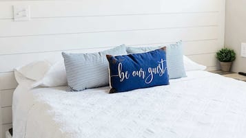 A bed with white bedding and a pillow with 'be our guest' text, a wall-mounted air conditioner, and a small potted plant.