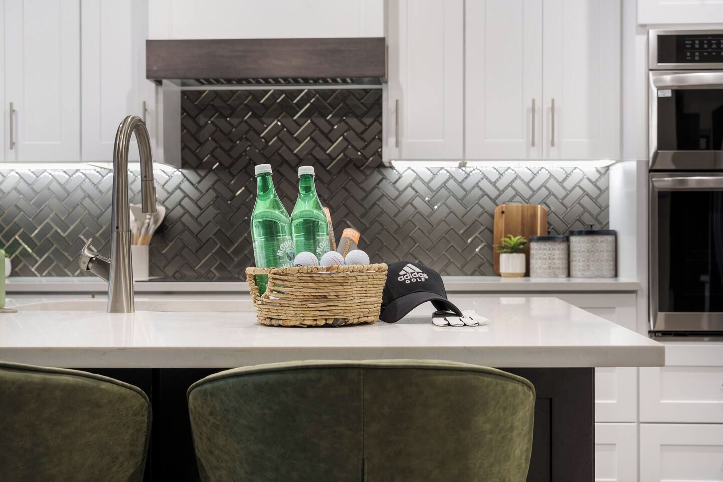 A kitchen with a stainless steel faucet, a herringbone-patterned backsplash, a stainless steel oven, and a woven basket containing bottles and a cap.