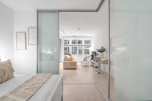 A modern bedroom with a glass partition, a bed with a textured blanket, and a view of a living area with a sofa and a TV. - Upscale Mercer Condo - Walk to Rogers Centre (Toronto)