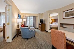 A hotel room with a bed, a desk, a chair, a television, and a window with curtains. - Accessible 2BR Suite w/ Tub | Breakfast + Pool (Kalamazoo)