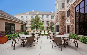 Un espace extérieur aménagé en terrasse, avec plusieurs tables et chaises, entouré de verdure et bordé d’un bâtiment à la façade en pierre.