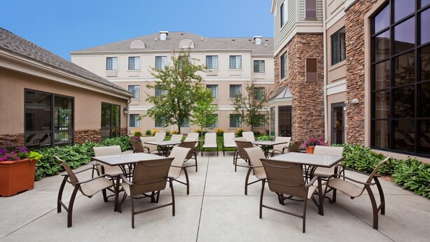 An outdoor patio area with multiple tables and chairs, surrounded by greenery and a building with a stone facade.