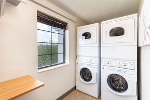 A laundry room with two stacked washing machines, a window with blinds, and a wooden bench.