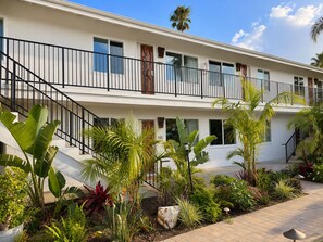 Un edificio de dos pisos con un balcón, rodeado de plantas tropicales y un camino pavimentado.