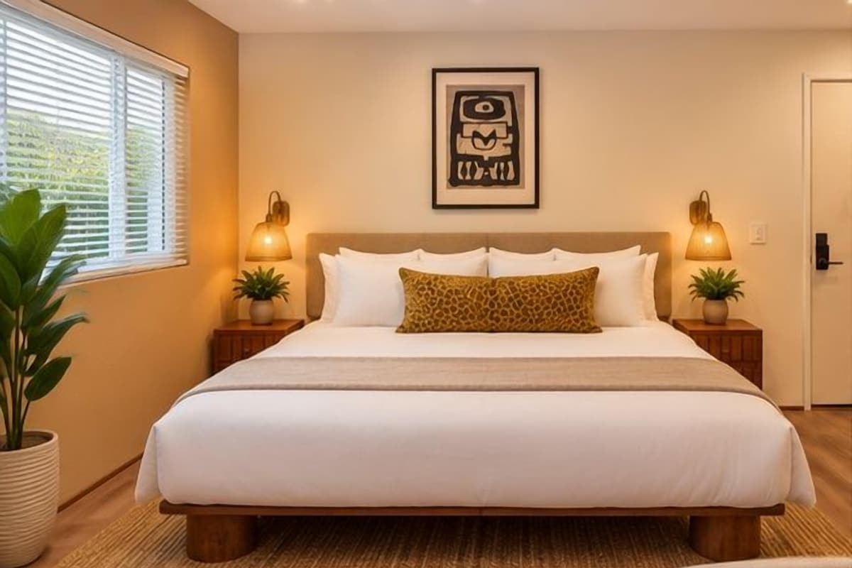 A bedroom with a large bed, two bedside lamps, a framed picture on the wall, and a window with blinds.