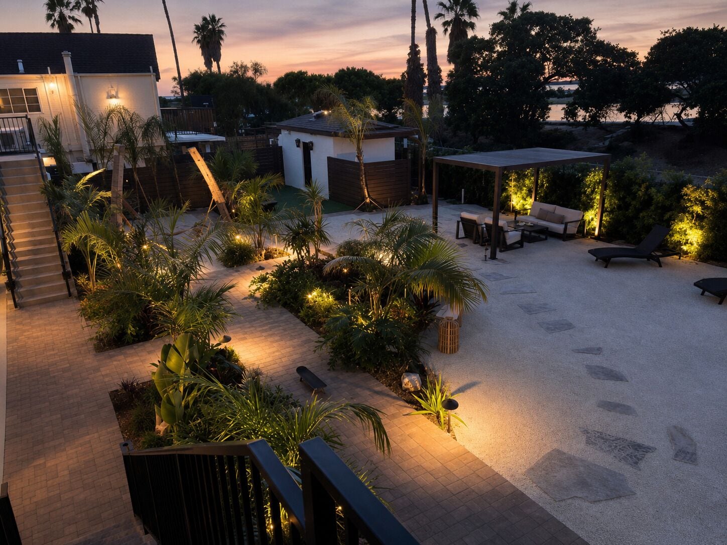 A well-lit outdoor patio area with a variety of seating options, including lounge chairs and a table, surrounded by lush greenery and palm trees.