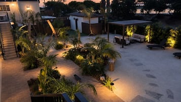 A well-lit outdoor patio area with a variety of seating options, including lounge chairs and a table, surrounded by lush greenery and palm trees.