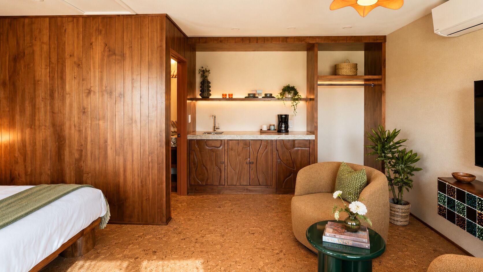 A room with a bed, a seating area with a table, a kitchen with wooden cabinets, and a TV mounted on the wall.