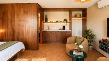A room with a bed, a seating area with a table, a kitchen with wooden cabinets, and a TV mounted on the wall.