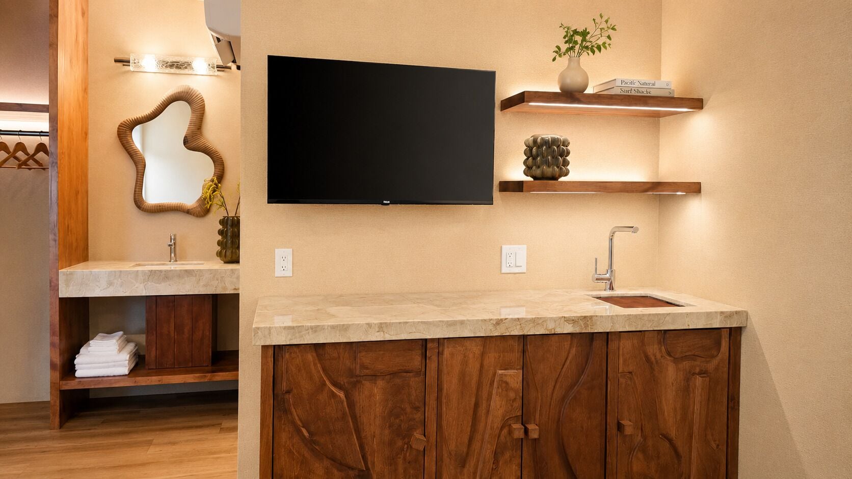 A modern bathroom with a wooden vanity, a large mirror, a flat-screen TV mounted on the wall, and built-in shelves with decorative items.