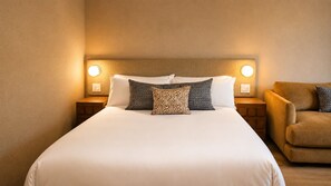Una cama bien hecha con cabecera, dos mesitas de noche con lámparas y un sillón marrón.