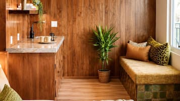 A compact room with a wooden wall, a small kitchenette, a seating area with cushions, a potted plant, and a window.