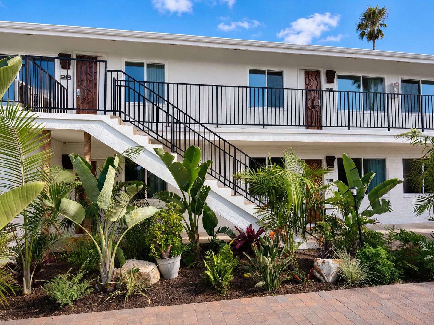A multi-story building with a white exterior, a staircase with black railings, and a variety of tropical plants in the foreground.