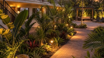 A hotel with a covered outdoor seating area, a paved walkway, and tropical landscaping.