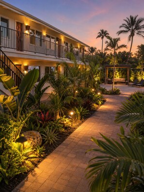 A hotel with a covered outdoor seating area, a paved walkway, and tropical landscaping.