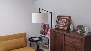 A room with a yellow sofa, a wooden cabinet with drawers, a small table with a lamp, and a bookshelf with various items. - Charming 1-bedroom bungalow with AC in Oak Bluffs (Oak Bluffs)