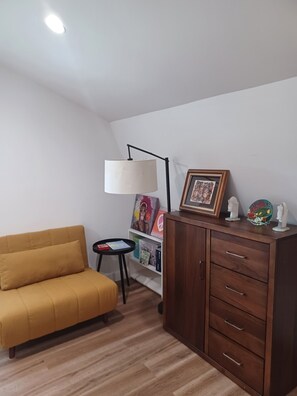 A room with a yellow sofa, a wooden cabinet with drawers, a small table with a lamp, and a bookshelf with various items. - Charming 1-bedroom bungalow with AC in Oak Bluffs (Oak Bluffs)