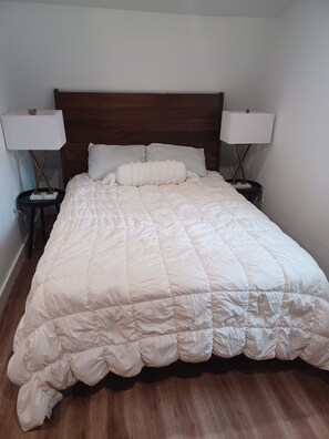 Uma cama bem arrumada com um edredom branco, duas mesas de cabeceira com abajures e uma cabeceira de madeira.