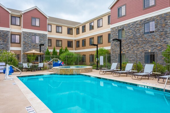 Una piscina con sillas de descanso y una zona de estar cubierta, rodeada por un edificio de varios pisos con fachada de piedra y madera.
