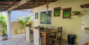 A covered patio with a wooden table and chairs, potted plants, and a window with a view of greenery. - Casa em Camacari com Piscina e Churrasqueira (Camaçari)