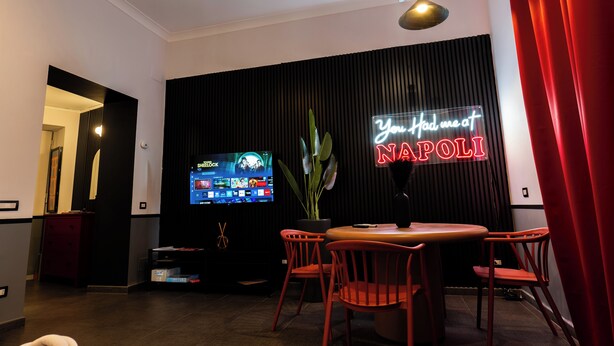 A modern living room with a flat-screen TV, a neon sign, a dining table, and red chairs. - LH51 Urban Suite – Design & Luxury in the Historic Center of Naples (Napoli)