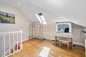 Ein Zimmer mit schrägen Wänden, einem Schminktisch mit Spiegel und Beleuchtung, einem Metallregal und einem Gemälde an der Wand.