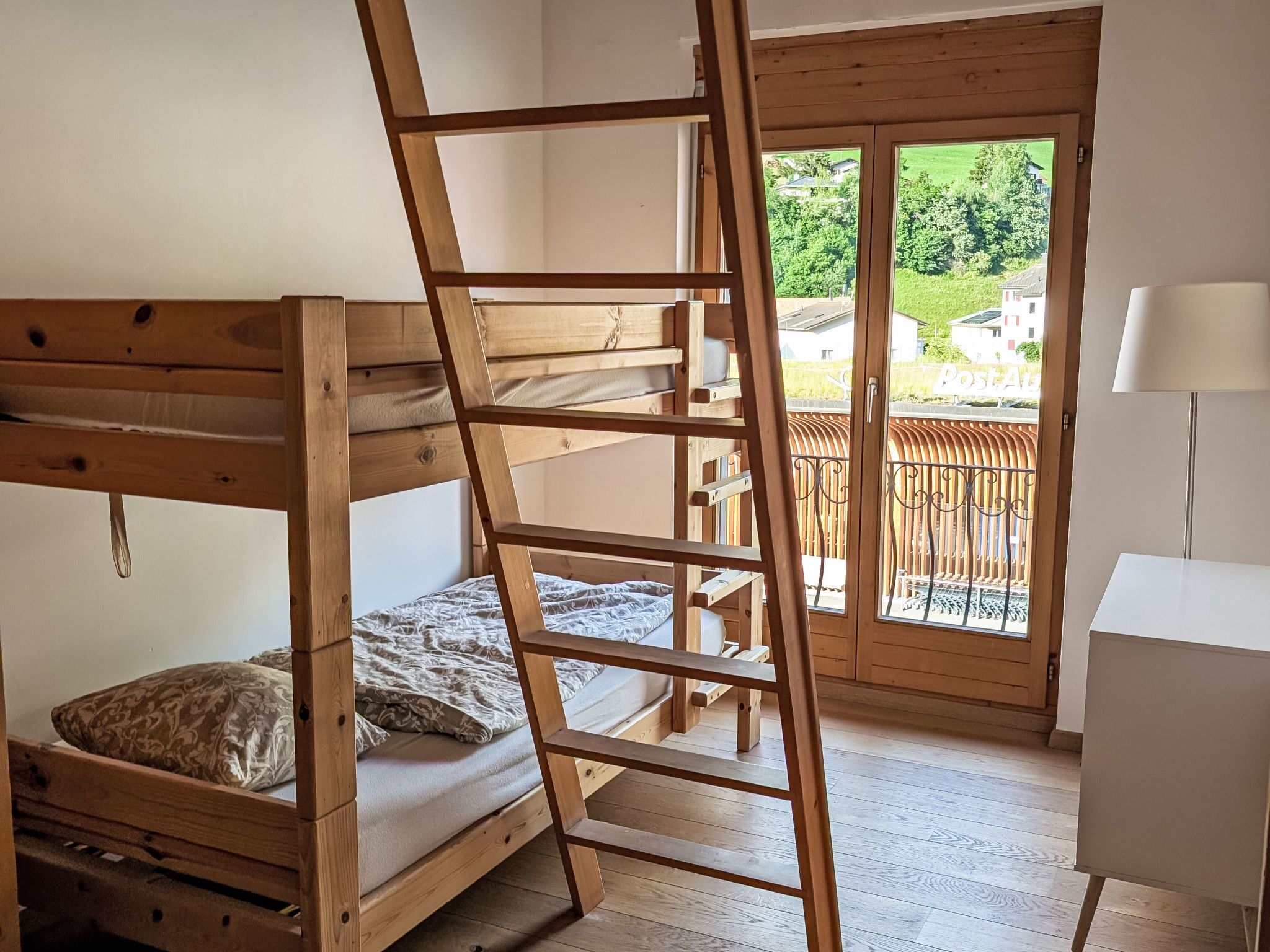A bunk bed with a wooden ladder, a white desk, and a view of a balcony with a fence.