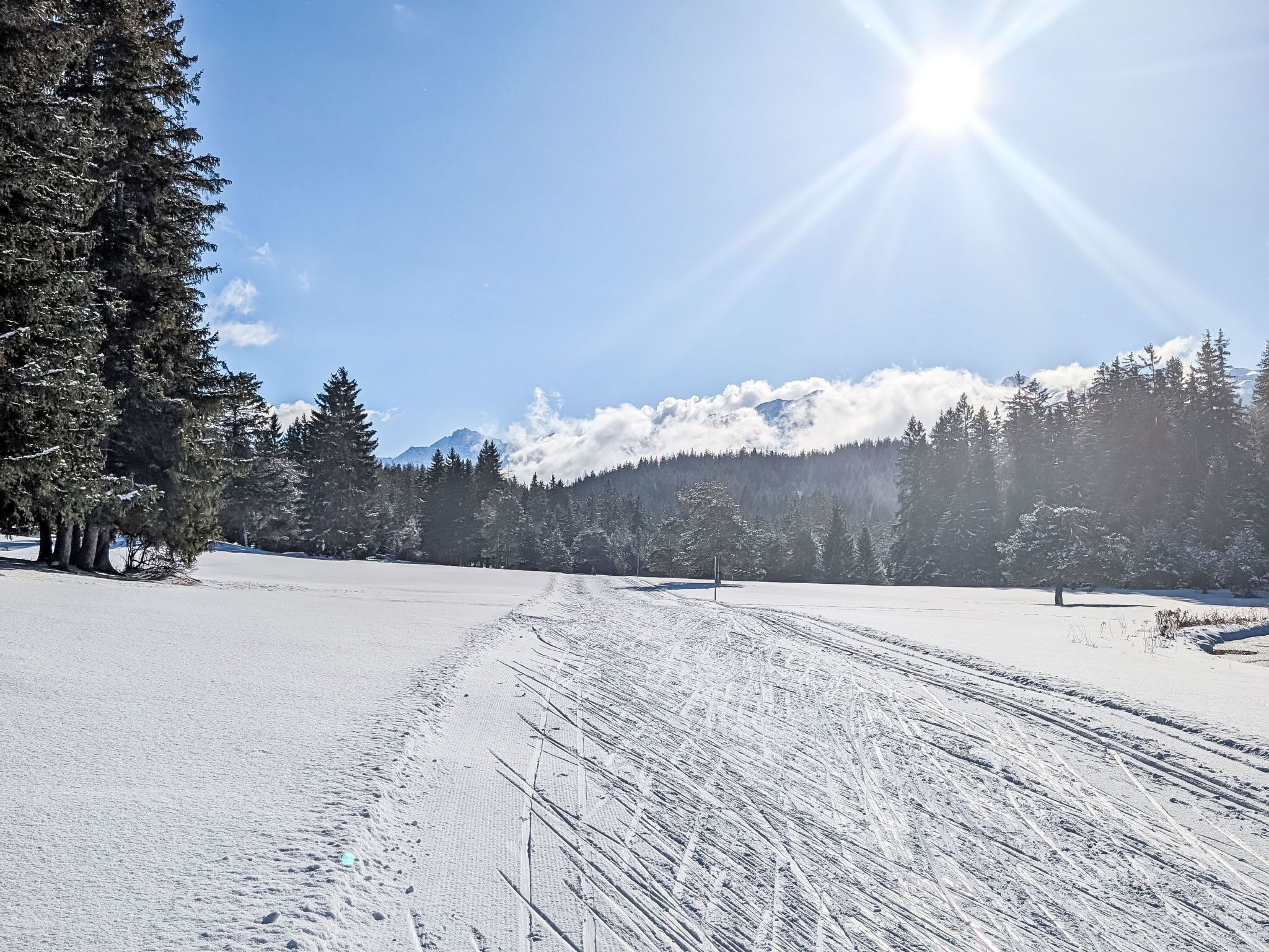 A snowy landscape with a clear sky, a sun shining, and a mountain range in the background.