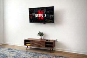 A wall-mounted television displaying the Netflix logo, a wooden console table with a plant, and a blue patterned rug. - Near Lake Texoma 1BR-Kingbed I Pool-GYM (Sherman)
