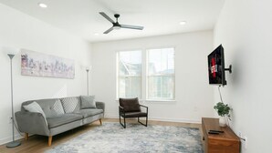 Sala de estar com sofá cinza, uma poltrona marrom, tapete azul e branco, ventilador de teto, TV montada na parede e uma pequena mesa lateral de madeira.
