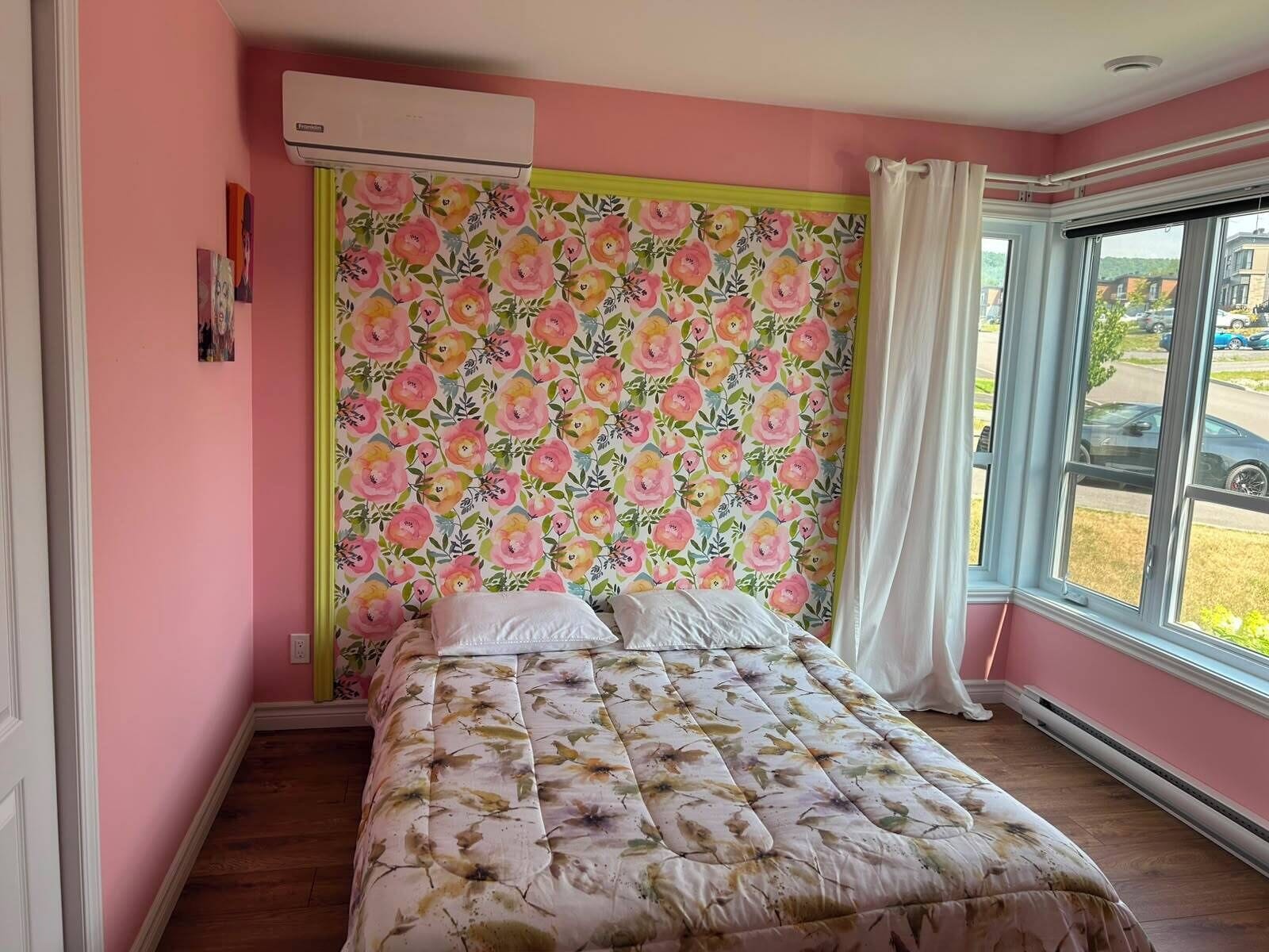 A bedroom with a floral-patterned headboard, a bed with a floral duvet, a window with a view of the outside, and an air conditioning unit on the wall.