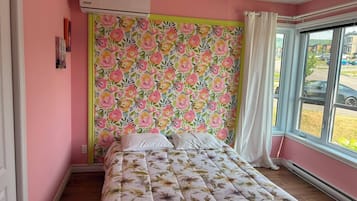 A bedroom with a floral-patterned headboard, a bed with a floral duvet, a window with a view of the outside, and an air conditioning unit on the wall.