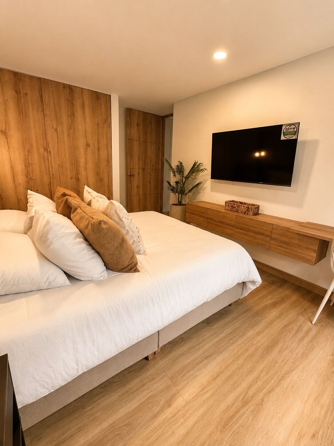 Une chambre à coucher moderne avec un grand lit, une tête de lit en bois, un téléviseur fixé au mur et une table de chevet en bois.
