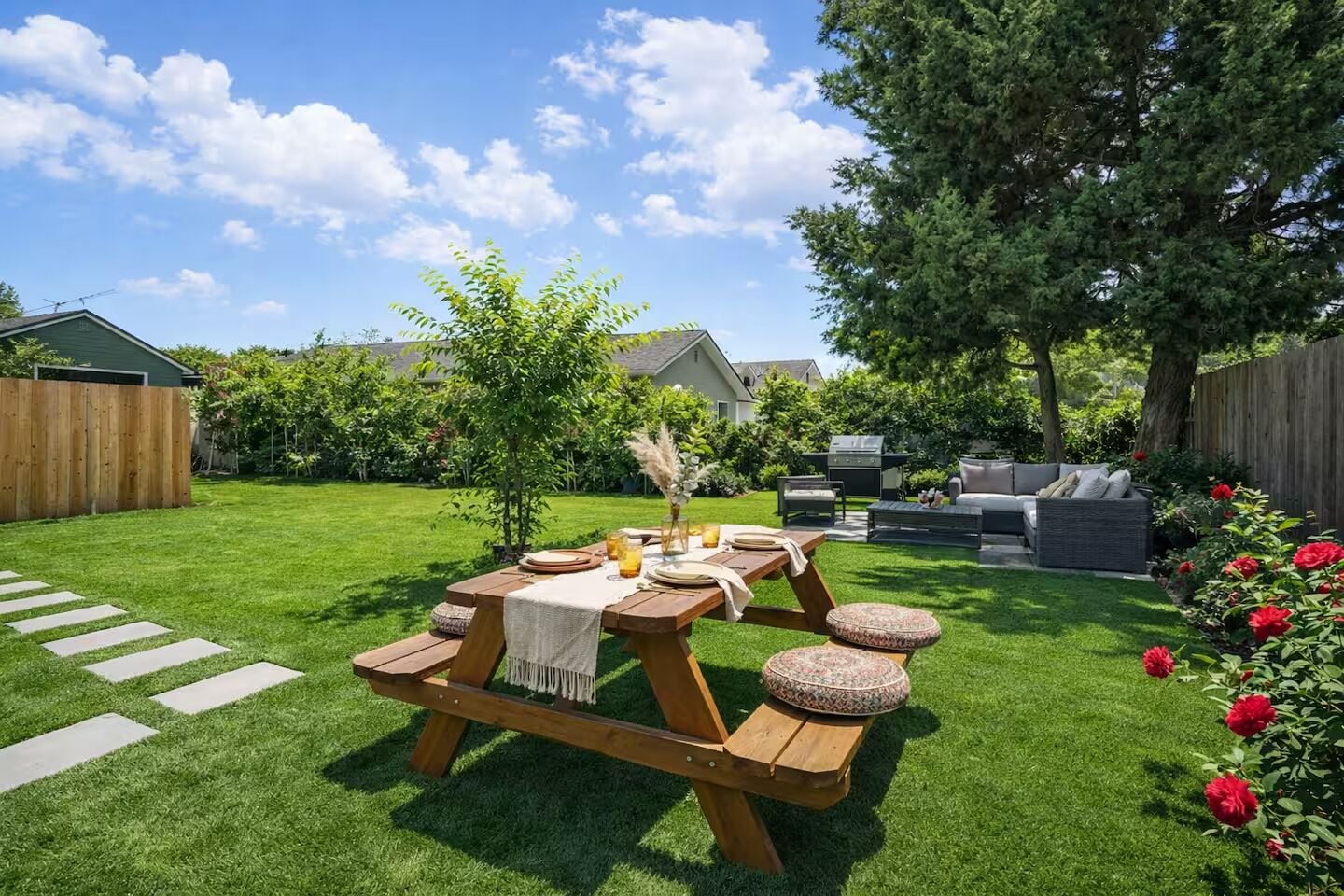A backyard with a wooden picnic table set for four, surrounded by lush greenery and a wooden fence.